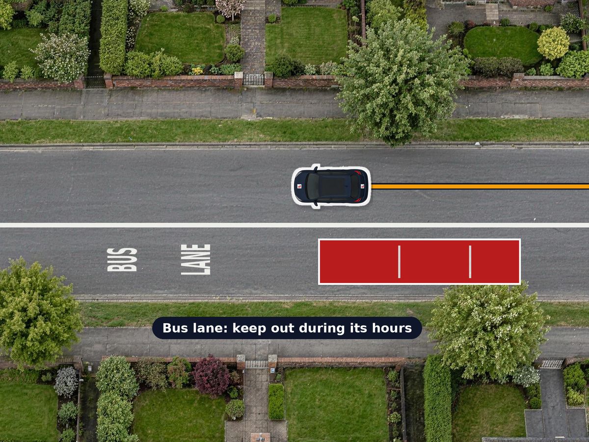 A one-way street from above. The left-hand lane is a bus lane, marked by a wide solid white line with BUS LANE painted in it and a bus driving in it. Your car keeps to the other lane.